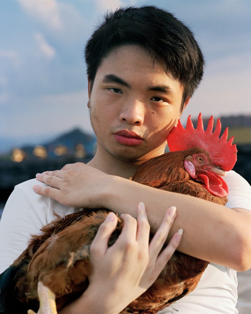 Person holding a red rooster, looking directly at the camera with a neutral expression.