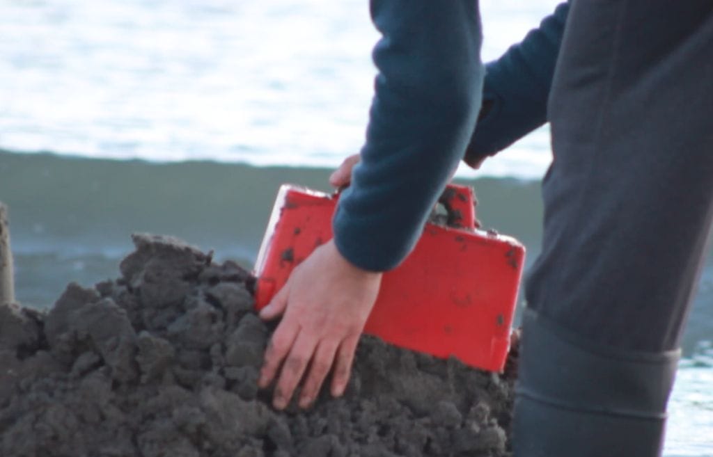 A person wearing dark sleeves shapes a mound of wet sand with a red rectangular mold, akin to a miniature Build a Wall Project, on the beach. The ocean is visible in the blurred background.