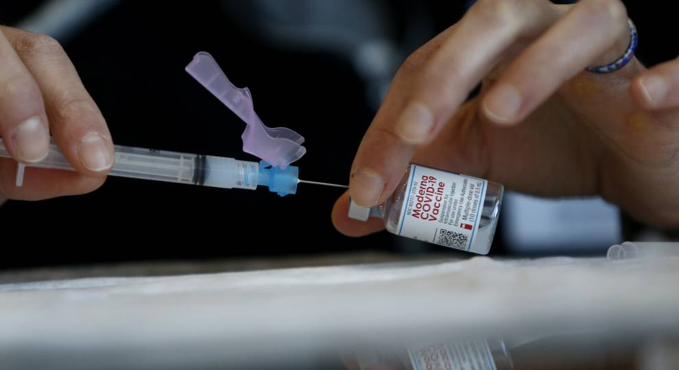 Hands hold a syringe and a vial labeled Moderna COVID-19 Vaccine against a blurred background, as if preparing for student vaccinations at colleges. One hand inserts the needle into the vial, while the other stabilizes it.
