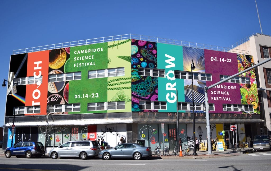 The image showcases a large, colorful mural on a building promoting the Cambridge Science Festival with the theme Touch, Grow. This vibrant example of environmental branding features scientific imagery and event dates 04.14-23.