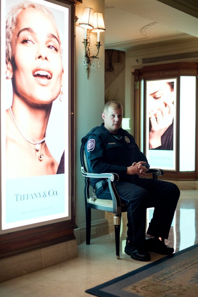 A security guard in uniform sits on a chair in a well-lit area, framed by lit Tiffany & Co. advertisement displays featuring models. The setting appears to be a hotel or a retail environment, with ornate lighting above.