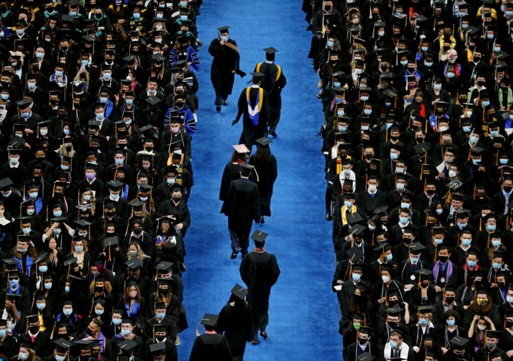 A large college commencement in Boston features rows of seated graduates in black gowns and caps. A blue carpeted aisle runs down the center, where more graduates from the of 2022 are walking. Many attendees are wearing face masks.
