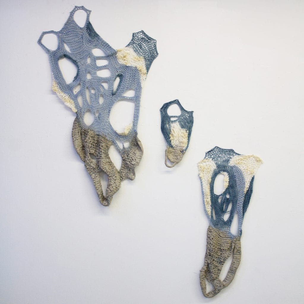 Abstract textile art with three irregularly shaped, interconnected pieces on a wall. The pieces are a mix of blue, beige, and white yarns with lace-like patterns, resembling organic forms. The largest piece is on the left, with two smaller ones nearby.