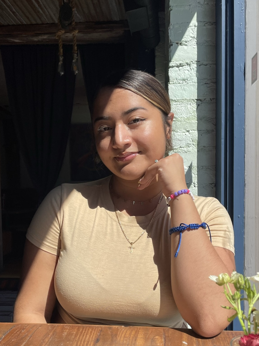 Woman in a beige top sits at a wooden table in a sunlit room, smiling slightly. She has her hand on her chin, wearing bracelets and a necklace, and is partially in shadow with sunlight illuminating part of her face.