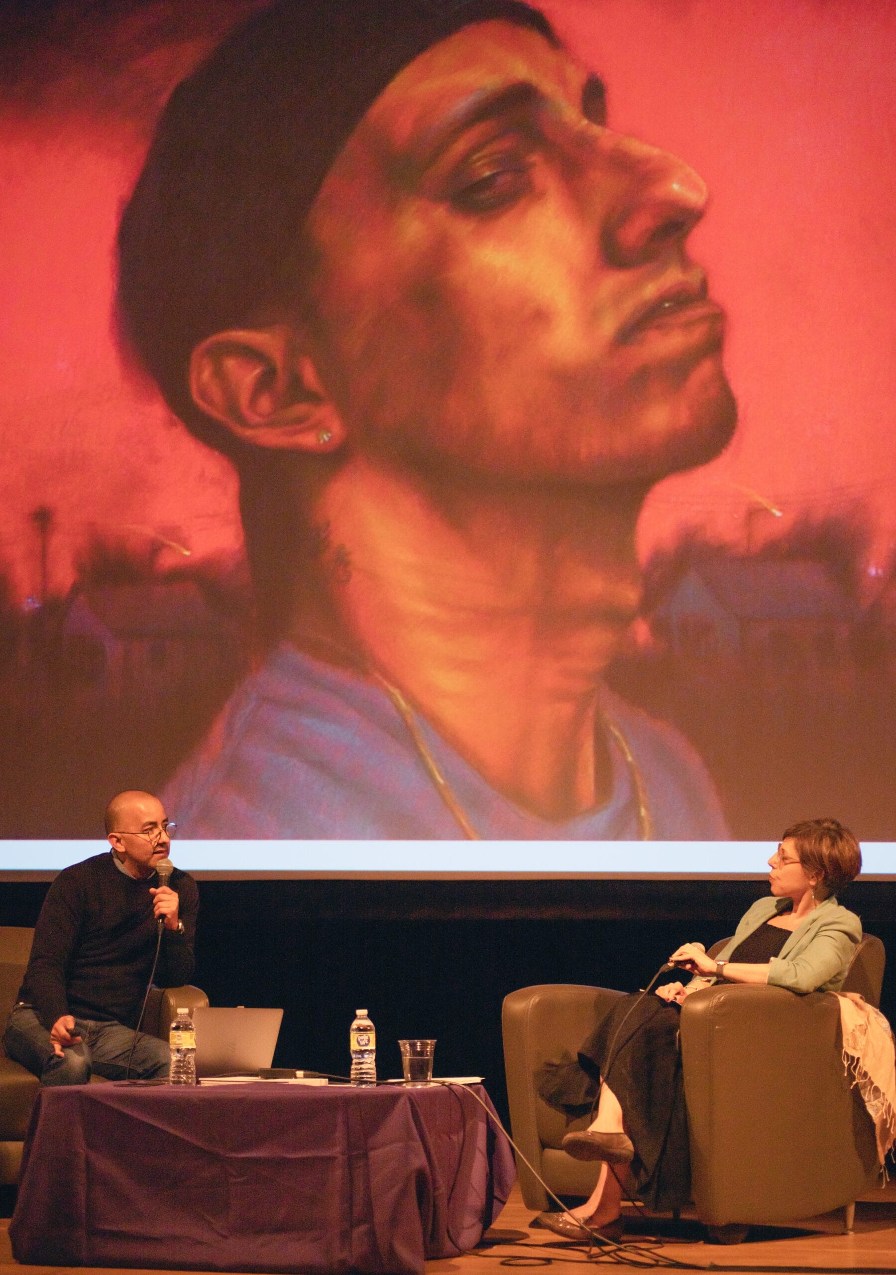 A man and woman on stage engage in a discussion while seated. Behind them is a large projected image of a painting: a close-up of a man with a serious expression, wearing a cap. A table with a laptop and water bottles is in the foreground.