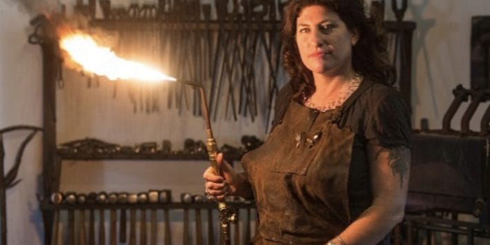In an industrial atmosphere, Corrina Sephora holds a lit torch in a workshop, clad in a protective apron. Various tools hang on the wall behind her as if each has its own story in Speaking of the Arts.