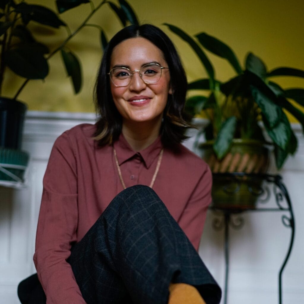 A person with glasses and shoulder-length hair, wearing a maroon shirt and plaid pants, sits on the floor smiling. They wear mustard yellow socks and white sneakers. Potted plants are in the background against a yellow wall.