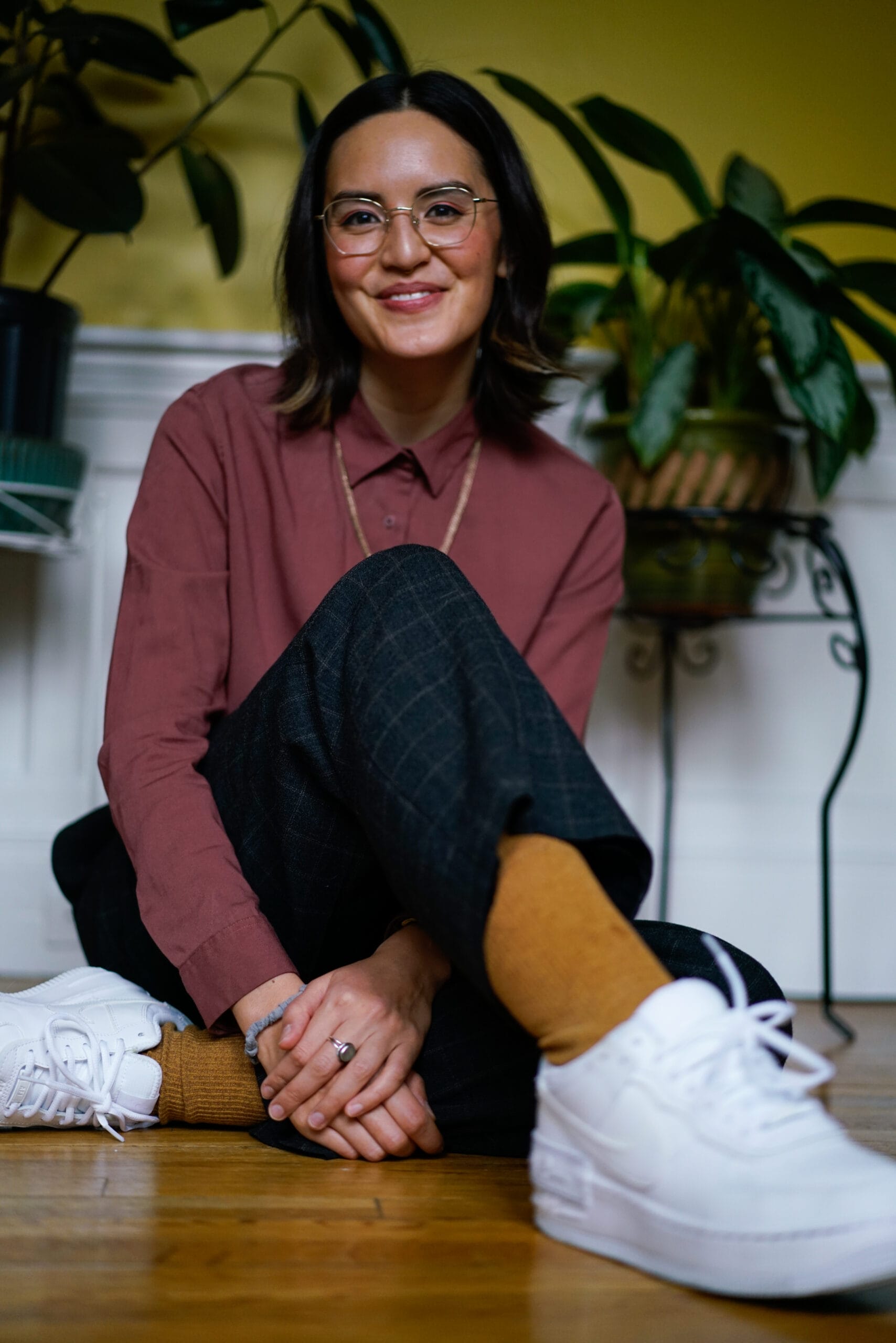 A person with glasses and shoulder-length hair, wearing a maroon shirt and plaid pants, sits on the floor smiling. They wear mustard yellow socks and white sneakers. Potted plants are in the background against a yellow wall.