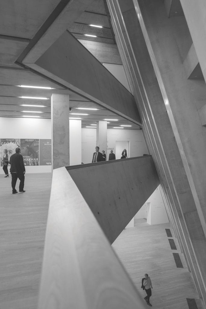 Black and white image of a modern, multi-level interior resembling the dynamic architecture of Londons Tate Modern. Concrete and wooden elements complement the geometric lines as people navigate different levels. Bright lighting enhances the minimalist design, evoking an art museum ambiance.