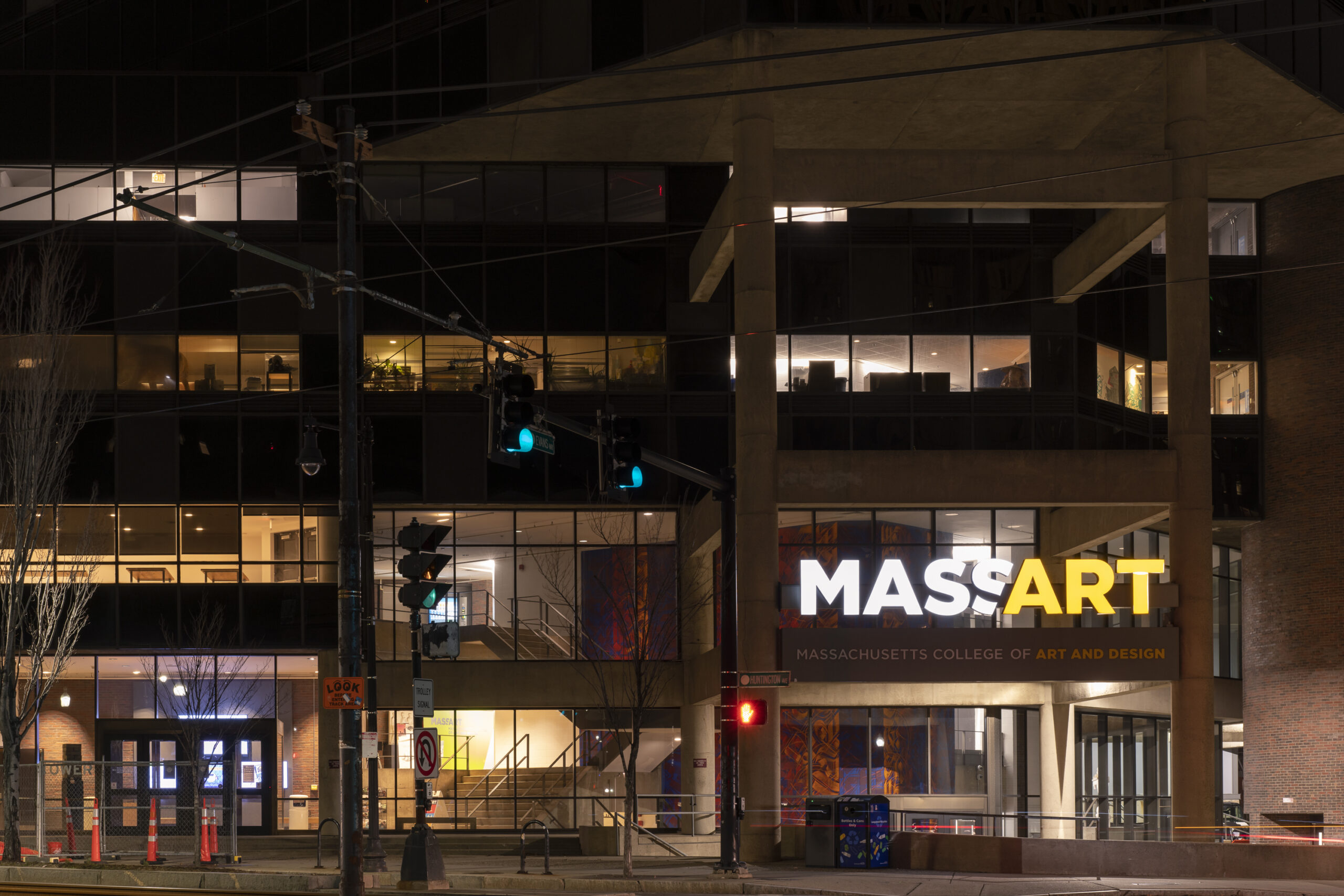 At night, the Massachusetts College of Art and Design building stands solemnly, its facade featuring large windows and a lit MASSART sign. Streetlights softly illuminate the quiet street in a poignant scene reminiscent of Boston Landmarks honoring COVID-19 victims.