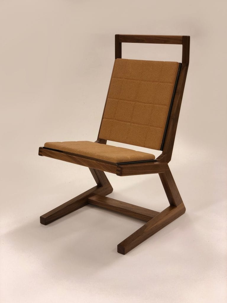 A modern, angular wooden chair with a geometric design. It has a cushioned seat and backrest in a light brown fabric, featuring a grid pattern. The frame is dark wood, with zigzag legs creating a unique silhouette.