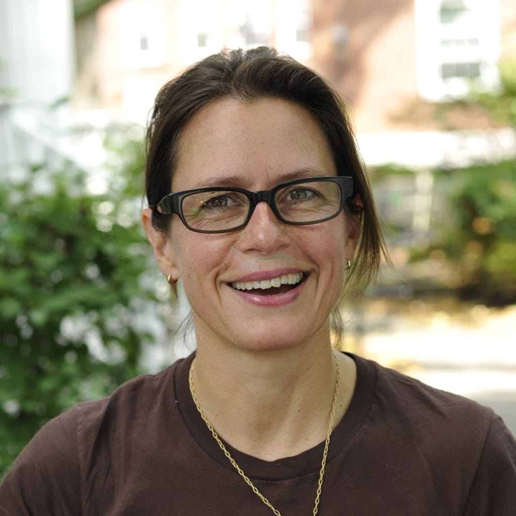 A person with brown hair and glasses is smiling outdoors. They are wearing a brown shirt and a gold necklace. The background is blurred with greenery and a building visible.