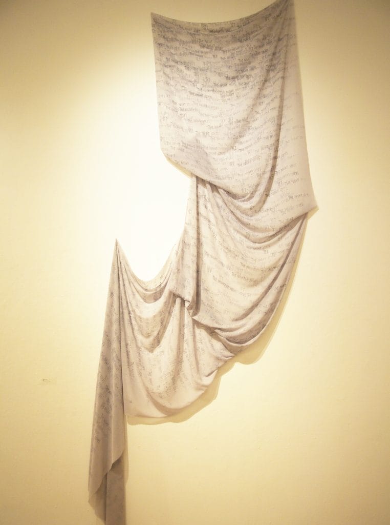A piece of fabric art draped against a beige wall. The light-colored fabric features handwritten text in a repeating pattern, creating a flowing, layered effect from top to bottom.