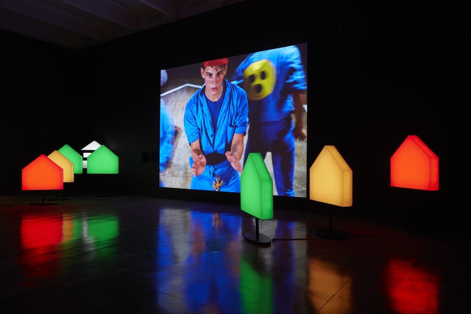 An art installation titled You The Better features a large video screen depicting a person in blue, surrounded by colorful, house-shaped light sculptures in red, green, and yellow. These cast vibrant reflections on a dimly lit floor for dramatic effect.