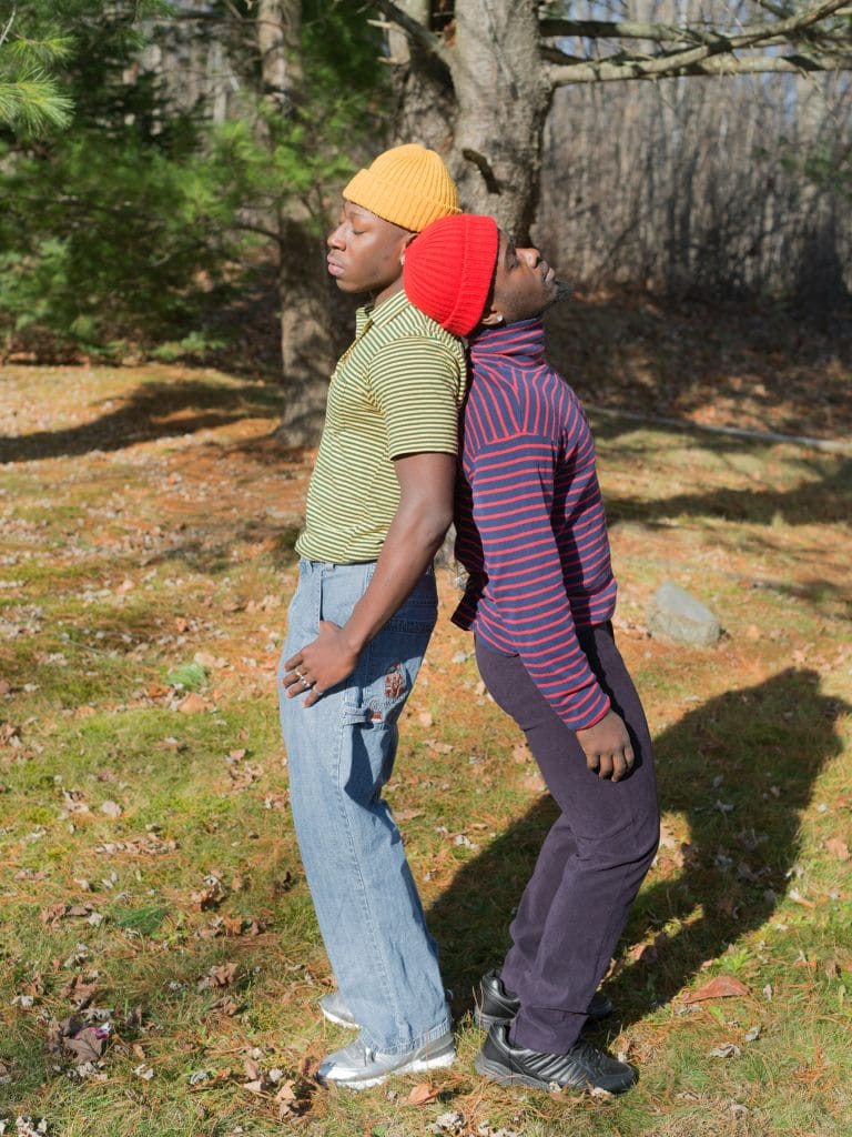 Two individuals, Uncle and Nana, stand back-to-back on a grassy area with scattered leaves, wearing colorful knit hats and striped shirts. One wears light blue jeans and the other dark pants. They appear relaxed, with trees and bare branches in the background.