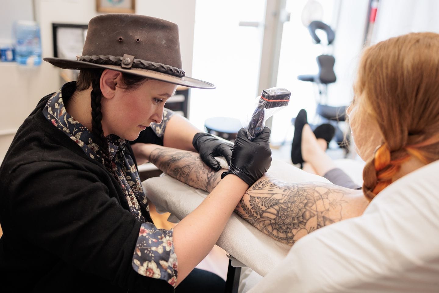 A tattoo artist in a hat and gloves is meticulously tattooing a floral design on the clients forearm, transforming skin into a canvas. The client, with long hair, reclines on a white chair amidst the studios array of tattoo equipment and art-adorned walls.