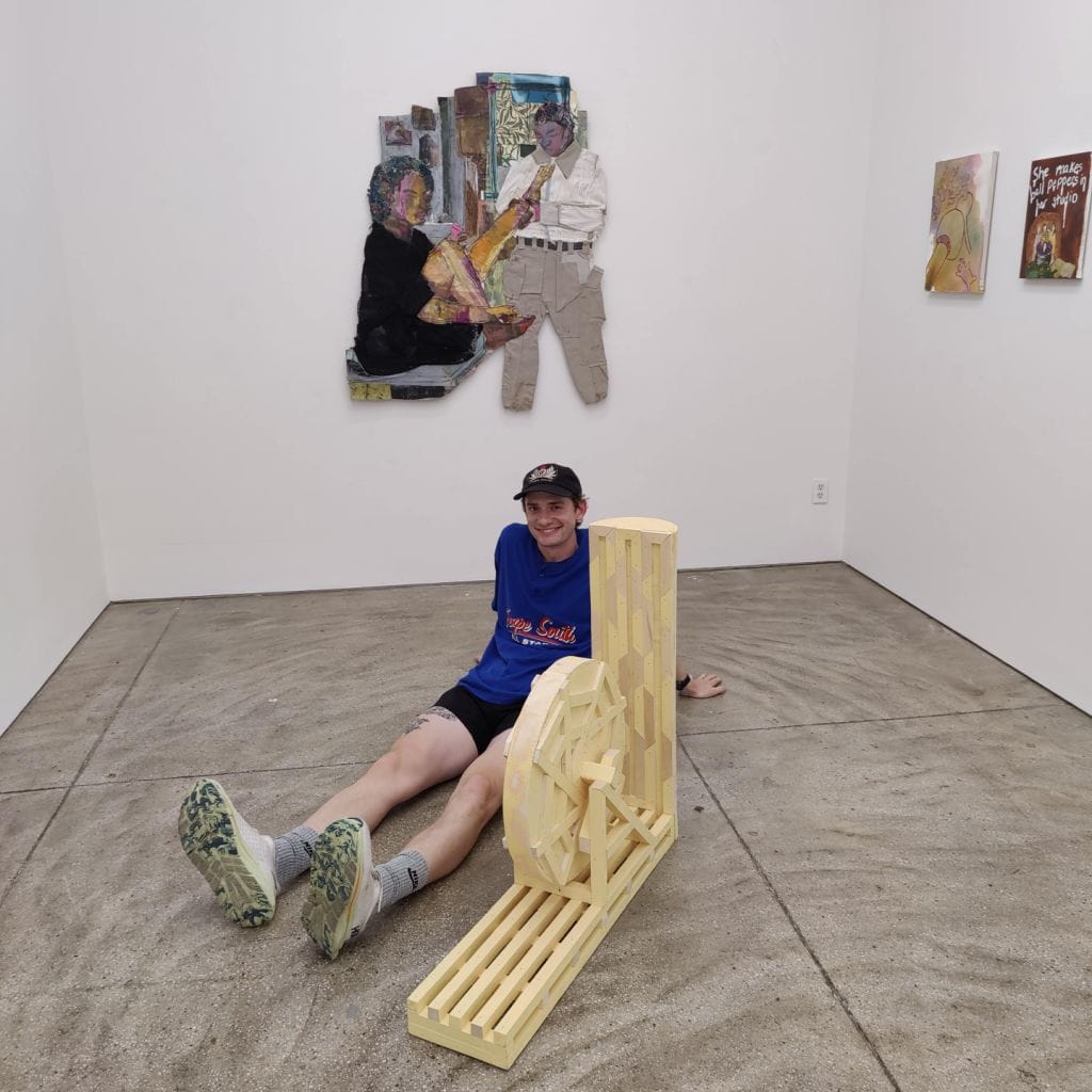 A person in a blue shirt and cap sits on a gallery floor, leaning against a sculptural piece made of wooden slats and a wheel. Behind them are several colorful mixed-media artworks on the white walls.