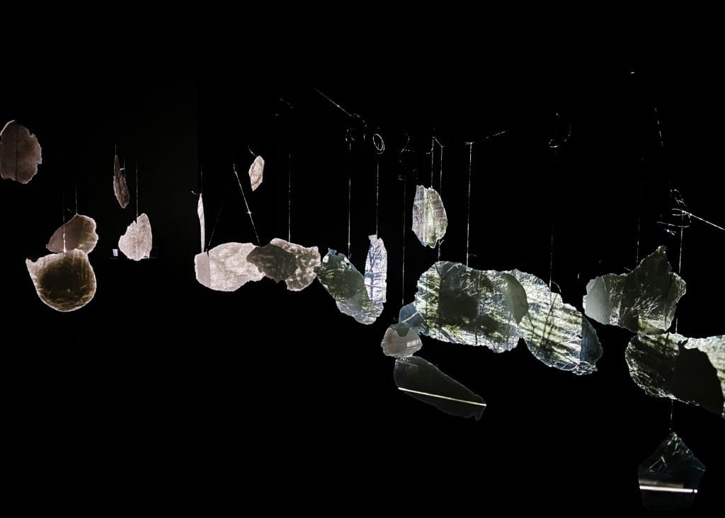 Translucent, irregularly-shaped pieces of material are suspended in the air against a black background. Light reflects through them, creating a shimmering effect.