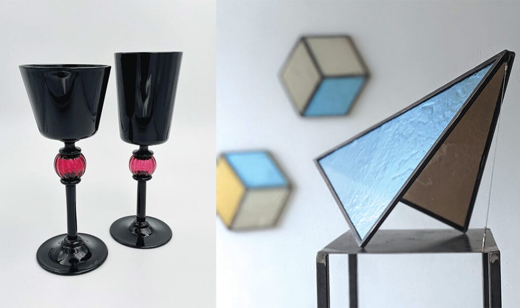 Two black wine glasses with pink accents on the left and a geometric sculpture featuring blue and bronze triangular panels on the right, are reminiscent of the stunning displays seen at the Janice Charach Gallery. Blurred cube shapes add depth to this complementary glass exhibit.