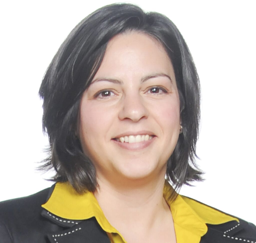 Smiling person with short dark hair, wearing a black blazer with white stitching and a bright yellow shirt underneath. The background is white.