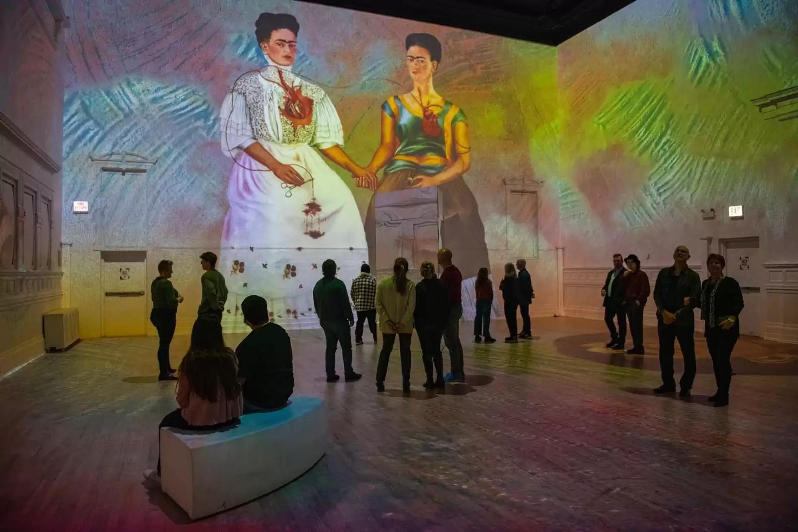 In a dimly lit room at a Boston museum, people observe a large, colorful projection of two women holding hands, reminiscent of Frida Kahlos art. The walls and floor reflect vibrant patterns, enhancing the immersive exhibit before it disappears.