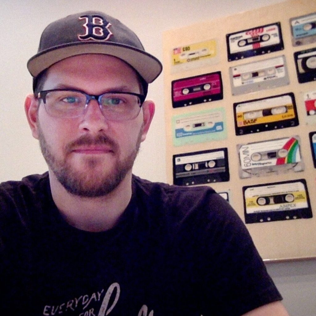 A person wearing glasses, a black T-shirt, and a baseball cap sits in front of a wall decorated with colorful cassette tapes.