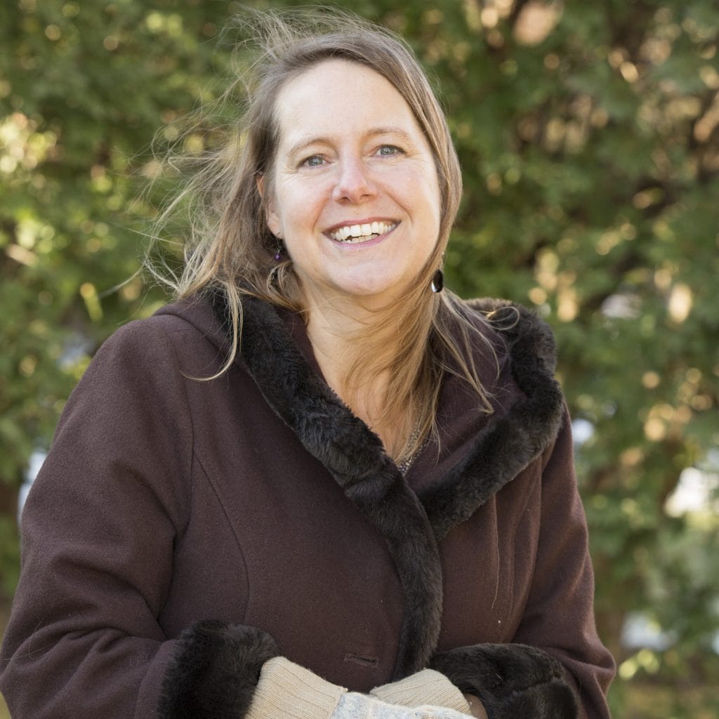 A woman with long hair smiling warmly, wearing a dark coat with a fur-trimmed collar and beige gloves. She stands outdoors with greenery in the background. The wind is gently blowing her hair.