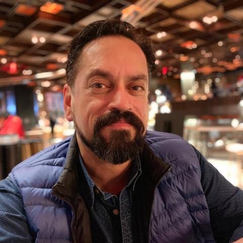 A person with a beard, wearing a blue puffer vest over a blue shirt, sits indoors at a restaurant with a warmly lit interior. The ceiling showcases a unique wooden design reminiscent of Fashion History elements that Jay Calderin might appreciate during his Last Call moments.