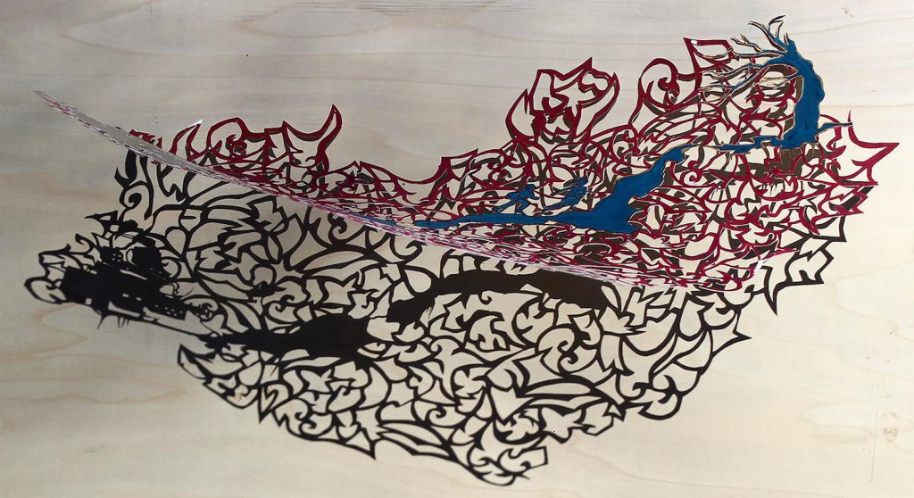Intricate cut-out sculpture with swirling, ornate patterns casting a detailed shadow on a wooden surface. The design incorporates red and blue elements, resembling an abstract, flowing organic form.