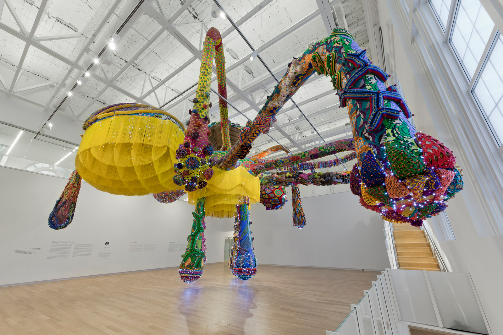 In the heart of MAAM, a large, colorful spider sculpture hangs from the gallery ceiling. This art-making marvel is intricately adorned with vibrant patterns, beads, and fabrics. Predominantly yellow with multicolored accents, it stands out against the white walls and wooden floor.