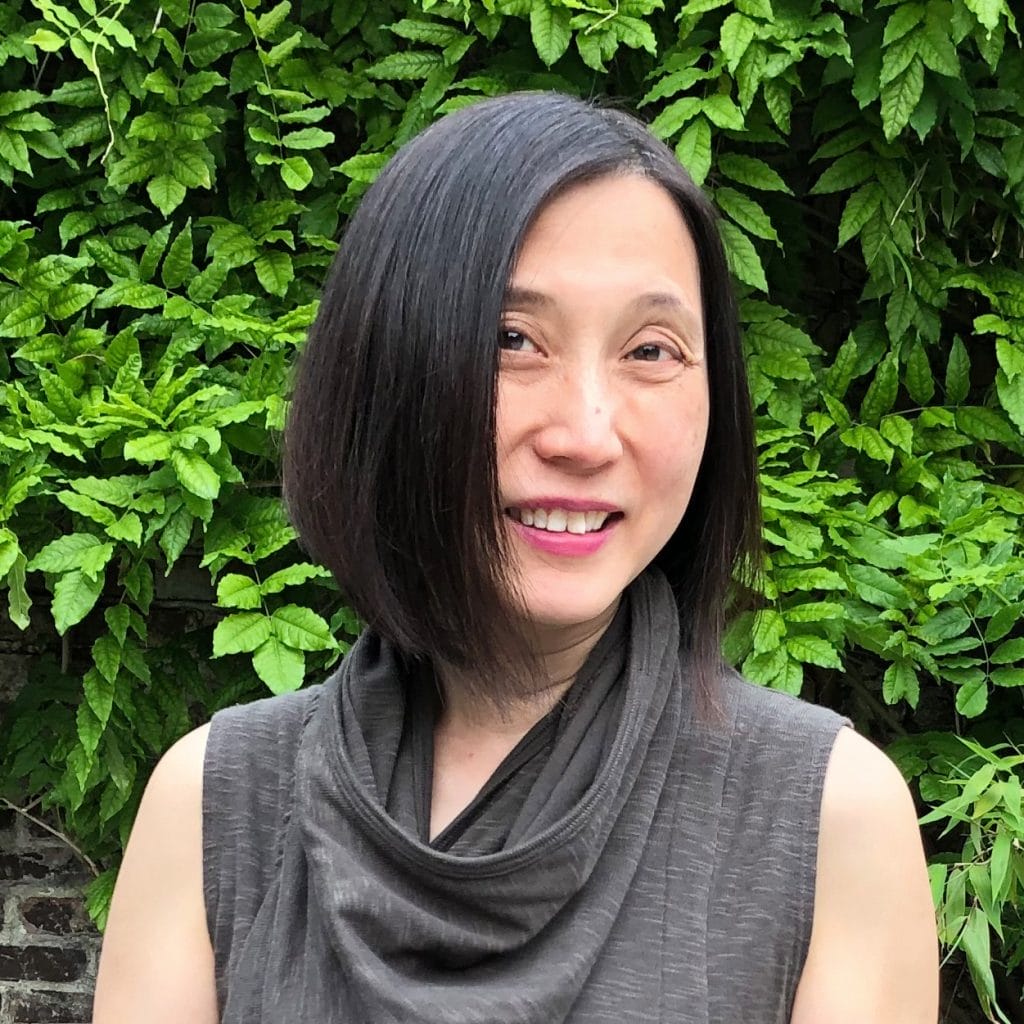 A person with a bob haircut, wearing a sleeveless dark top, smiles against a backdrop of lush green leaves.