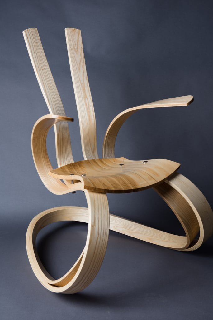 A modern wooden chair with a unique design featuring flowing, curved lines and smooth, natural woodgrain. The Ribbon Rocker boasts an artistic appearance with exaggerated, looping armrests and legs against a dark gray background.