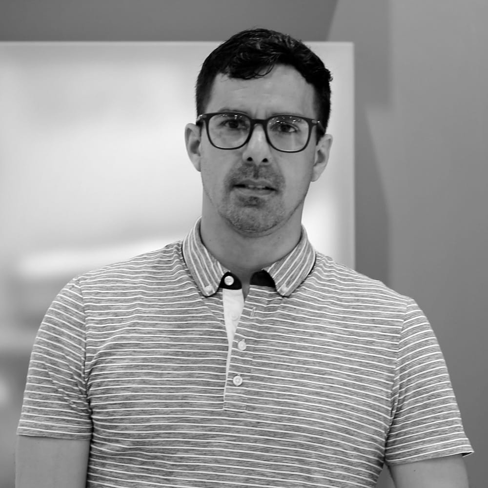 A black and white photo of a person wearing glasses and a striped polo shirt, standing indoors with a blurred, light-colored background.