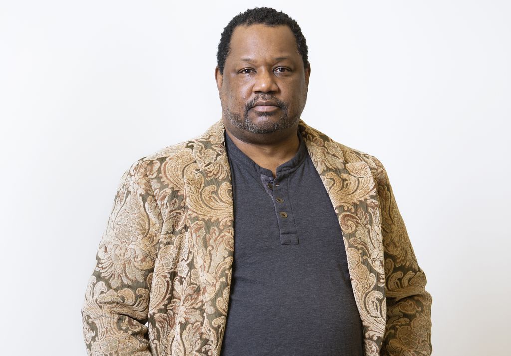 Man wearing a patterned jacket and dark shirt stands against a plain white background, looking confidently at the camera, channeling an air of creativity reminiscent of artists featured at the deCordova Sculpture Park and Museum.