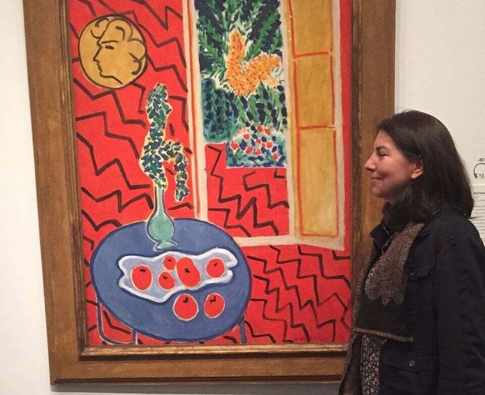 A person stands in profile, smiling at a vibrant painting in a gallery. The artwork features a red zigzag background, a window view, potted plant, and a dish of red fruit on a blue table.