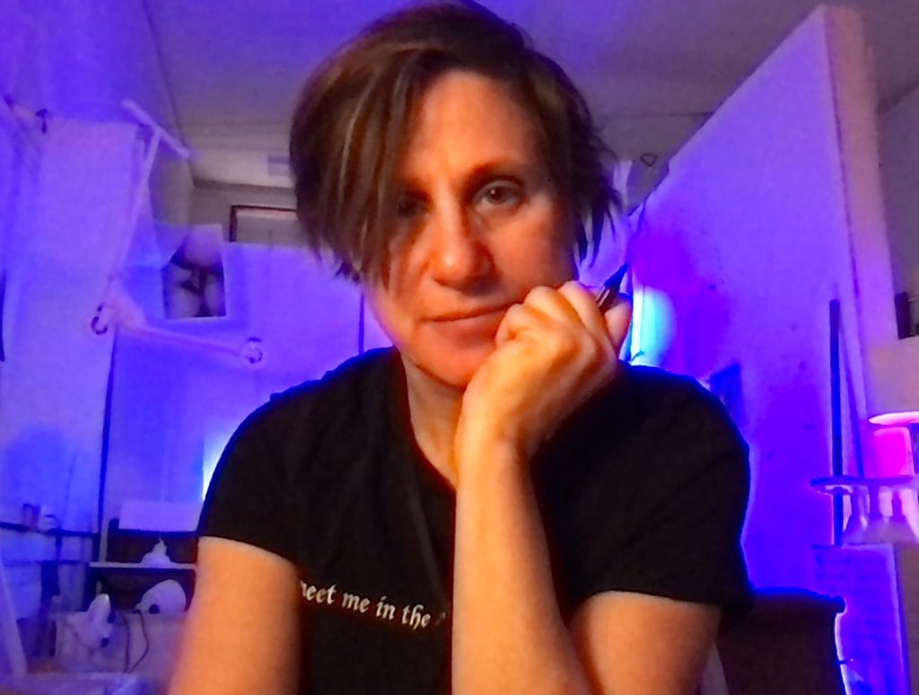 Person with short hair and a pensive expression, resting their chin on their hand. They are wearing a black T-shirt in a room lit with purple and blue lights. The background includes shelving and various items.