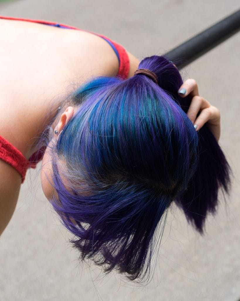 Jayne, with vibrant blue and purple hair tied in a ponytail, leans forward over a black bar. They wear a red top, their nails are perfectly painted. The blurred background highlights the striking hues in Jaynes hair and the elegant curve of their upper back.