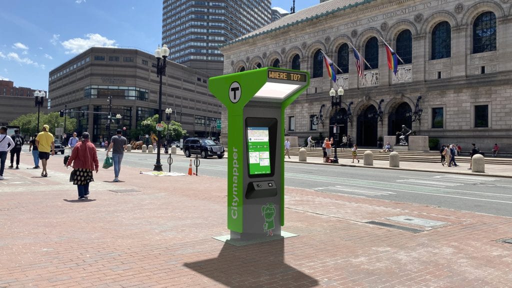An outdoor Citymapper kiosk stands in a bustling city plaza with its screen asking, Where to? It sits near a historic building as people stroll by, with streets and modern architecture forming the backdrop under a clear blue sky.