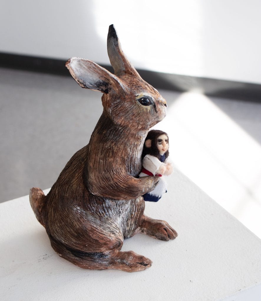 A sculpture of a large brown rabbit sitting and holding a small human figure. The rabbit has detailed fur texture and the human figure is wearing white and dark clothing. The sculpture is placed on a white pedestal in a well-lit room.