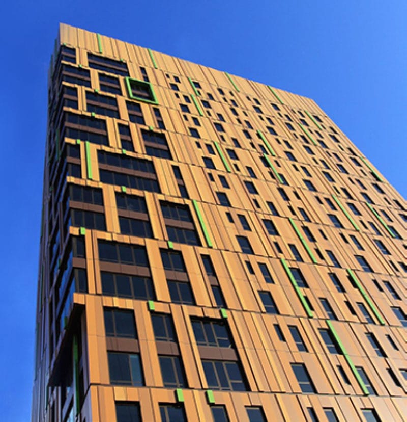 The tall modern building boasts a distinctive facade of orange and green vertical panels crafted by MCM Fabricators, complemented by large windows that glisten against the clear blue sky.