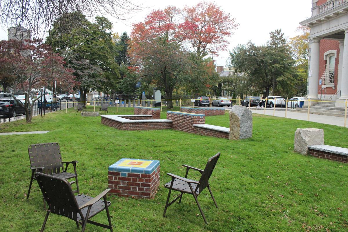 Four chairs encircle a brick table adorned with tile art on a grassy area in Jamaica Plain. Nearby, brick benches and large stones offer additional seating. Trees in autumn foliage border the area, creating a scenic setting for the public artwork, with a building and parked cars beyond.