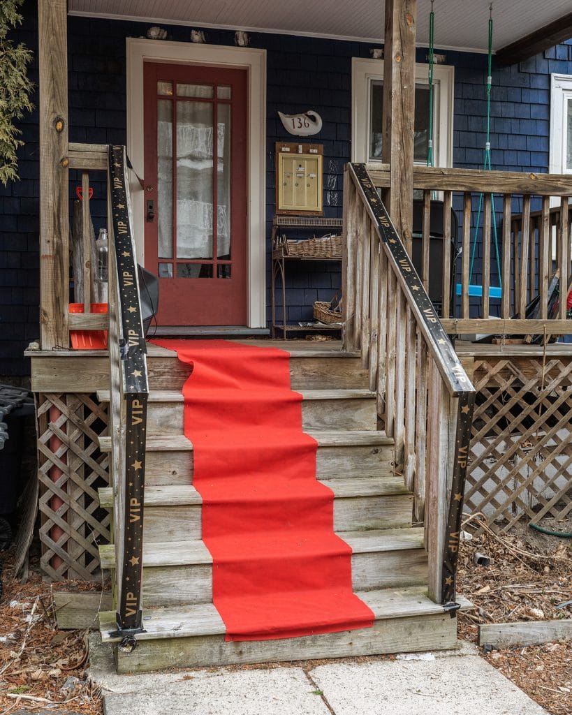 A wooden staircase leads up to a porch with a red carpet along the steps. The porch has a black railing decorated with VIP tape. The house exterior is blue with a red door. The number 136 is displayed above the mailboxes.