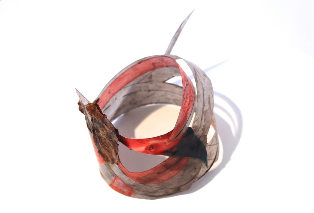 A spiraling abstract sculpture with a mix of translucent red and gray materials, featuring pointed extensions. The piece casts shadows on a white surface, creating a dynamic interplay of light and form.