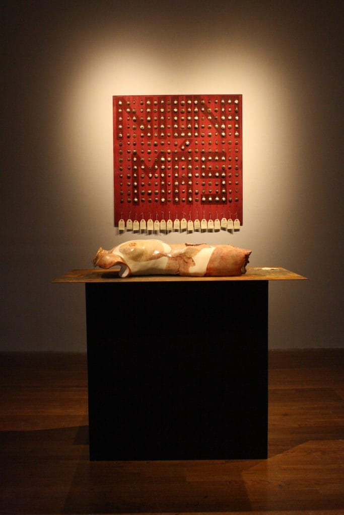 An art installation presents a human figure sculpture on a black pedestal. Behind it, a red panel emblazoned with NI UNA MÁS and circular patterns invites viewers to confront reality, all softly illuminated by a spotlight.