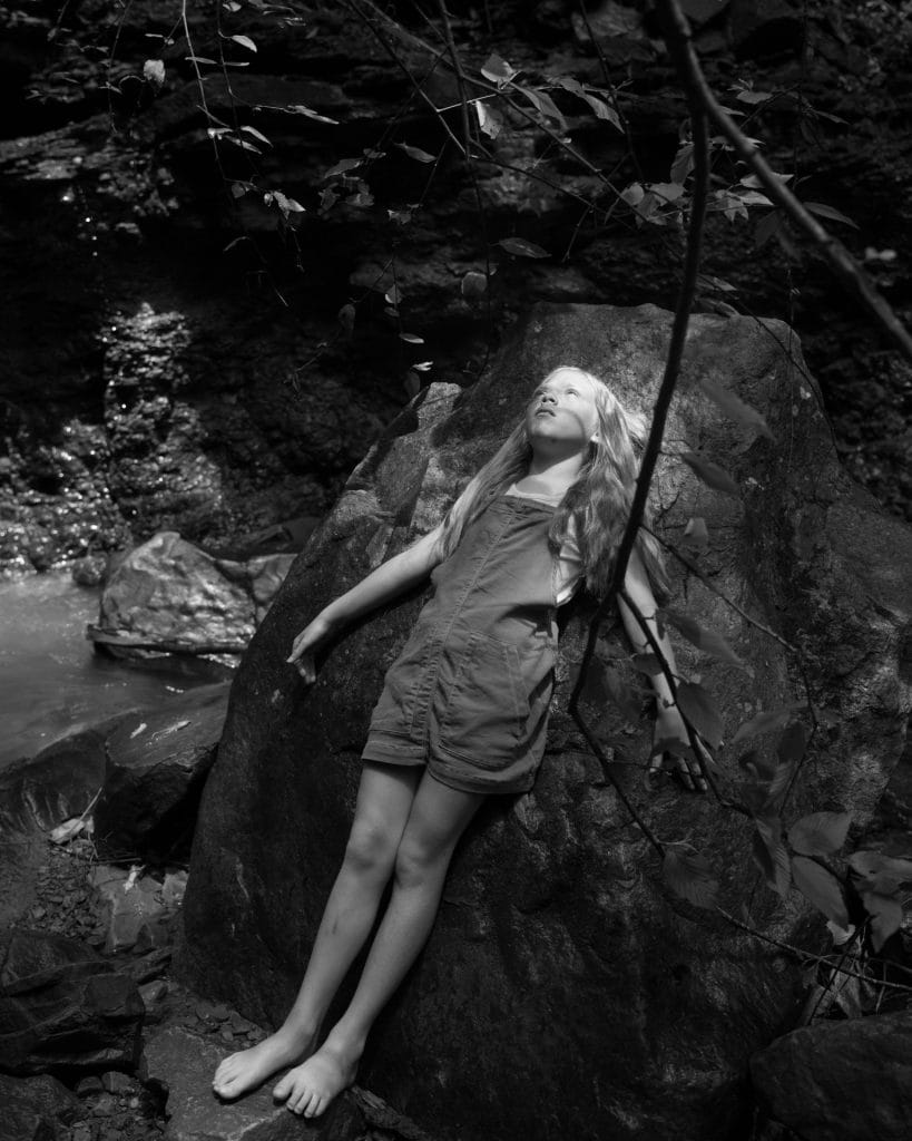 A barefoot child with long hair, wearing overalls, leans against a large rock in a forested area. Sunlight filters through the trees above, illuminating their face. The background has dark foliage and a small waterfall.