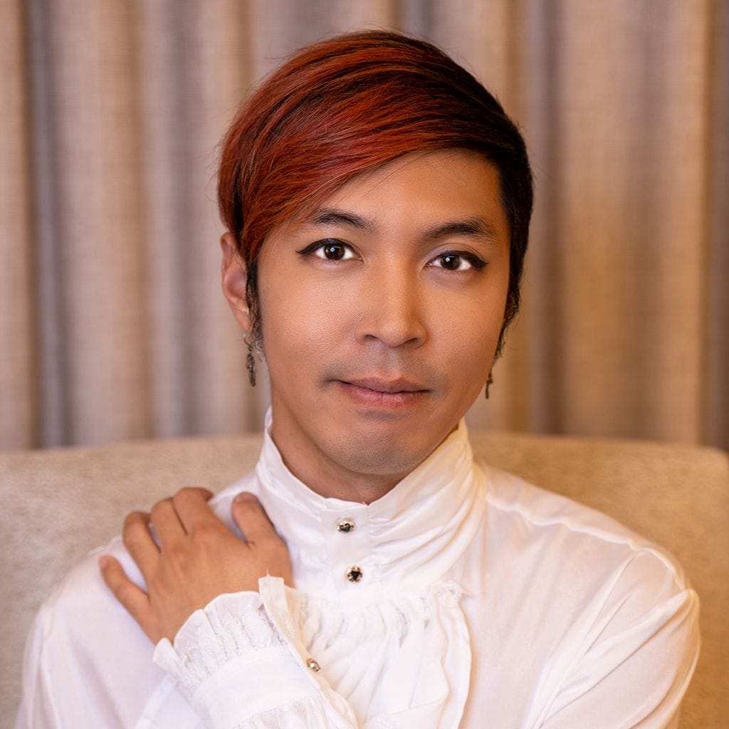 A person with short red hair and eyeliner is wearing a white ruffled shirt with decorative buttons. They have a gentle expression, with one hand placed on their shoulder. The background is a softly blurred curtain.