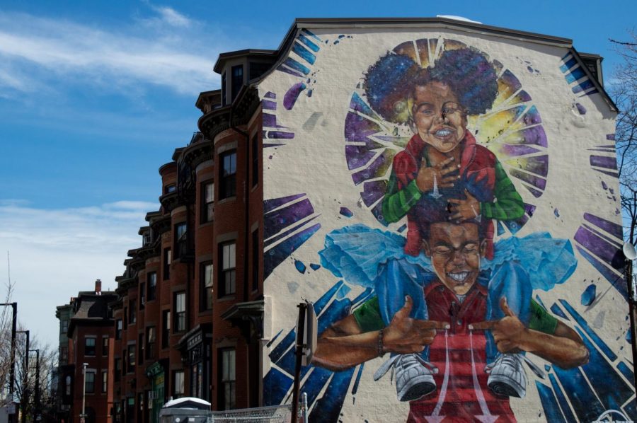 A vibrant mural on a building in an urban area with historic architecture depicts a smiling child sitting on an adults shoulders. With colors bursting against a clear blue sky, this artwork, inspired by Boston Creatives, embodies radical imagination and dreams of racial justice.