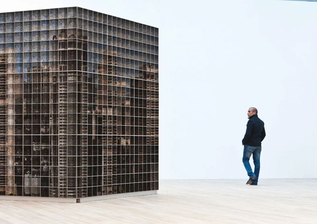 A person in a black jacket and jeans observes a large, grid-like sculpture resembling a building facade in an art gallery. The room has light wooden flooring and white walls, creating a minimalist setting.