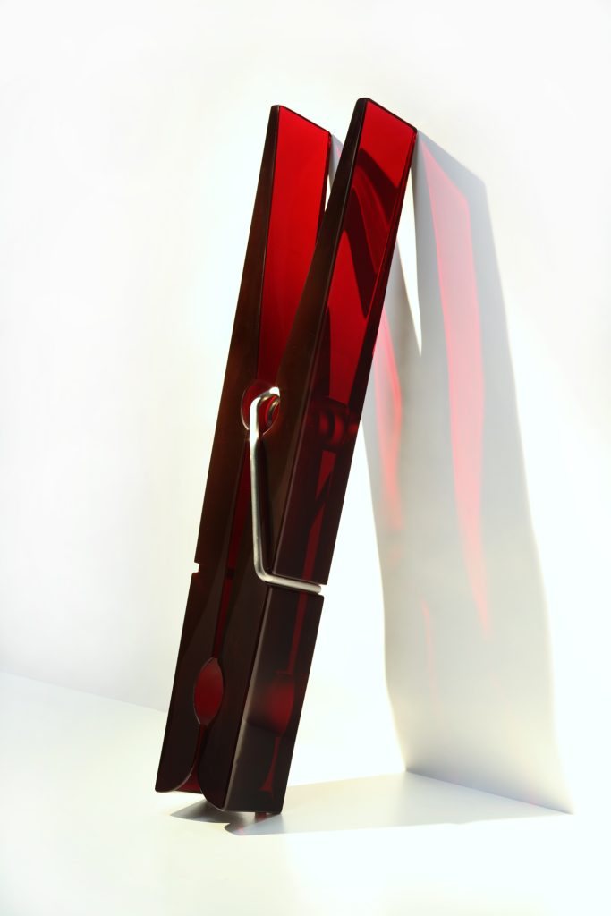A large red clothespin sculpture stands upright against a white background. The light casts a shadow, emphasizing the modern, minimalist design and glossy finish of the red sculpture.
