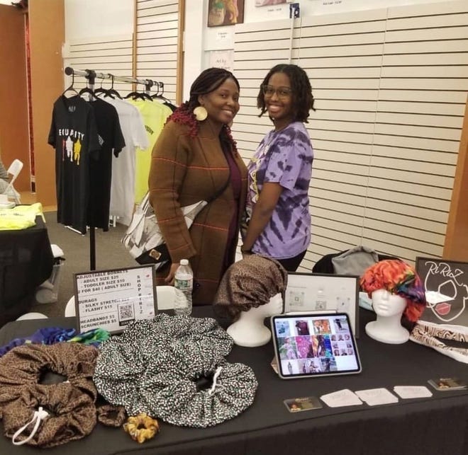 Two people stand smiling behind a display table at the Holiday Market in Brockton. The table features colorful patterned bonnets, fabric scrunchies, and a tablet with images. Behind them are clothing racks with various garments, capturing the essence of Small Business Saturday.